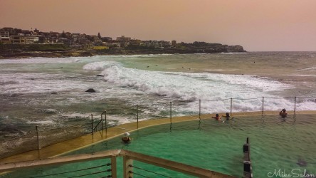 Swimmers wading in the clear water of Bronte Ocean Pool while large waves crash against the outer sea wall under a hazy morning sky.